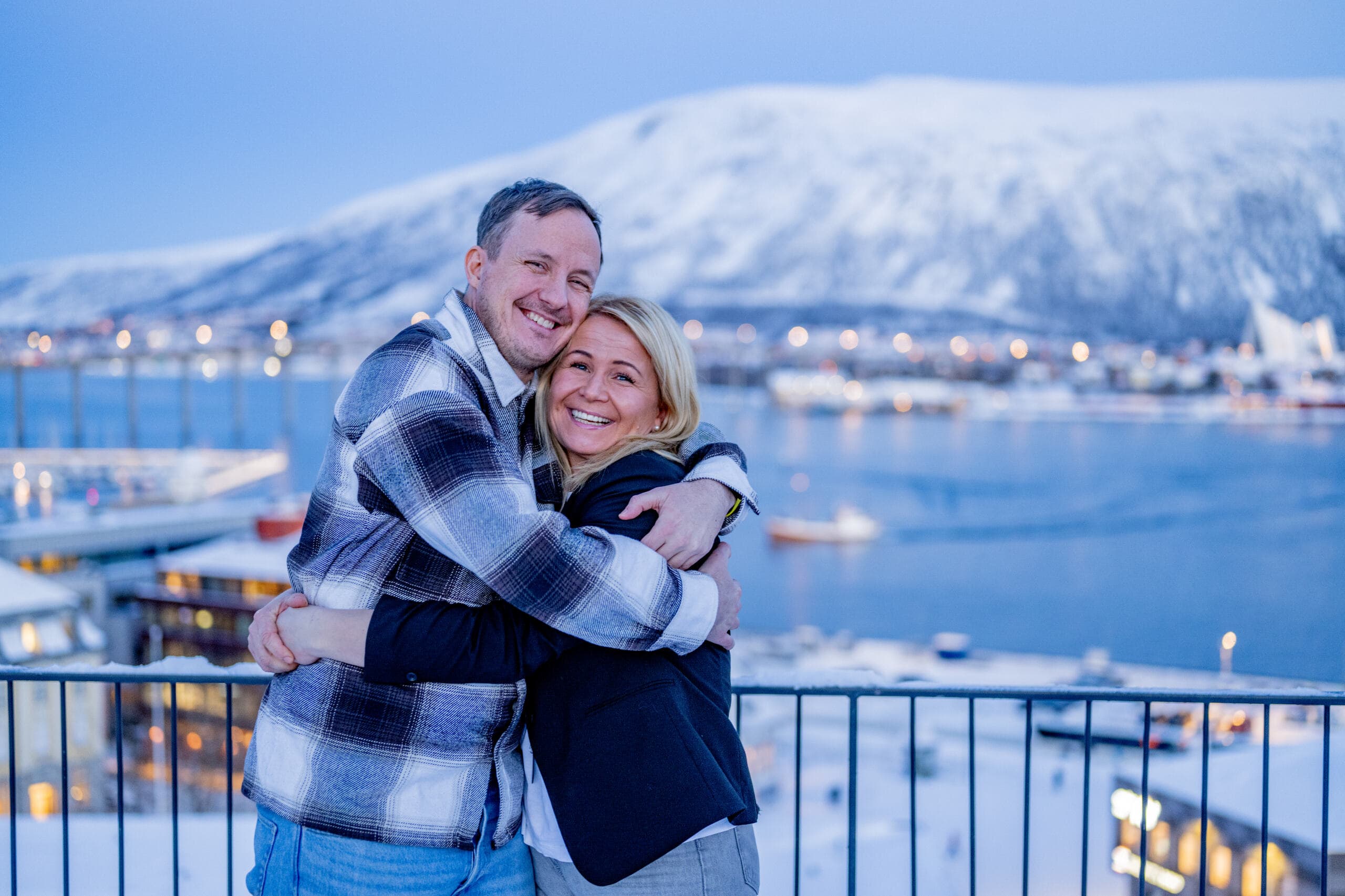 Et smilende par klemmer hverandre på en snødekt balkong med utsikt over en naturskjønn havnefront med fjell i bakgrunnen om vinteren.
