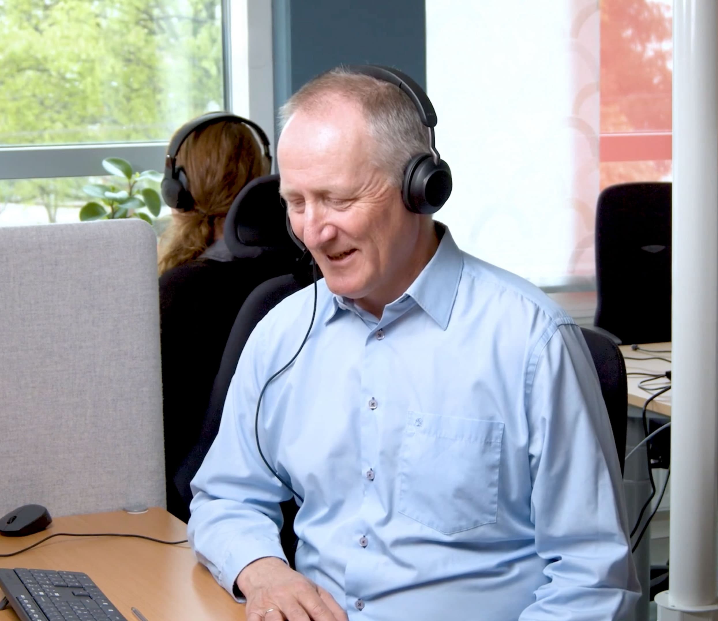En mann i lyseblå skjorte og headset sitter ved et skrivebord og smiler mens han arbeider på en datamaskin på et kontor. En annen person med hodesett sitter ved en arbeidsstasjon i nærheten.
