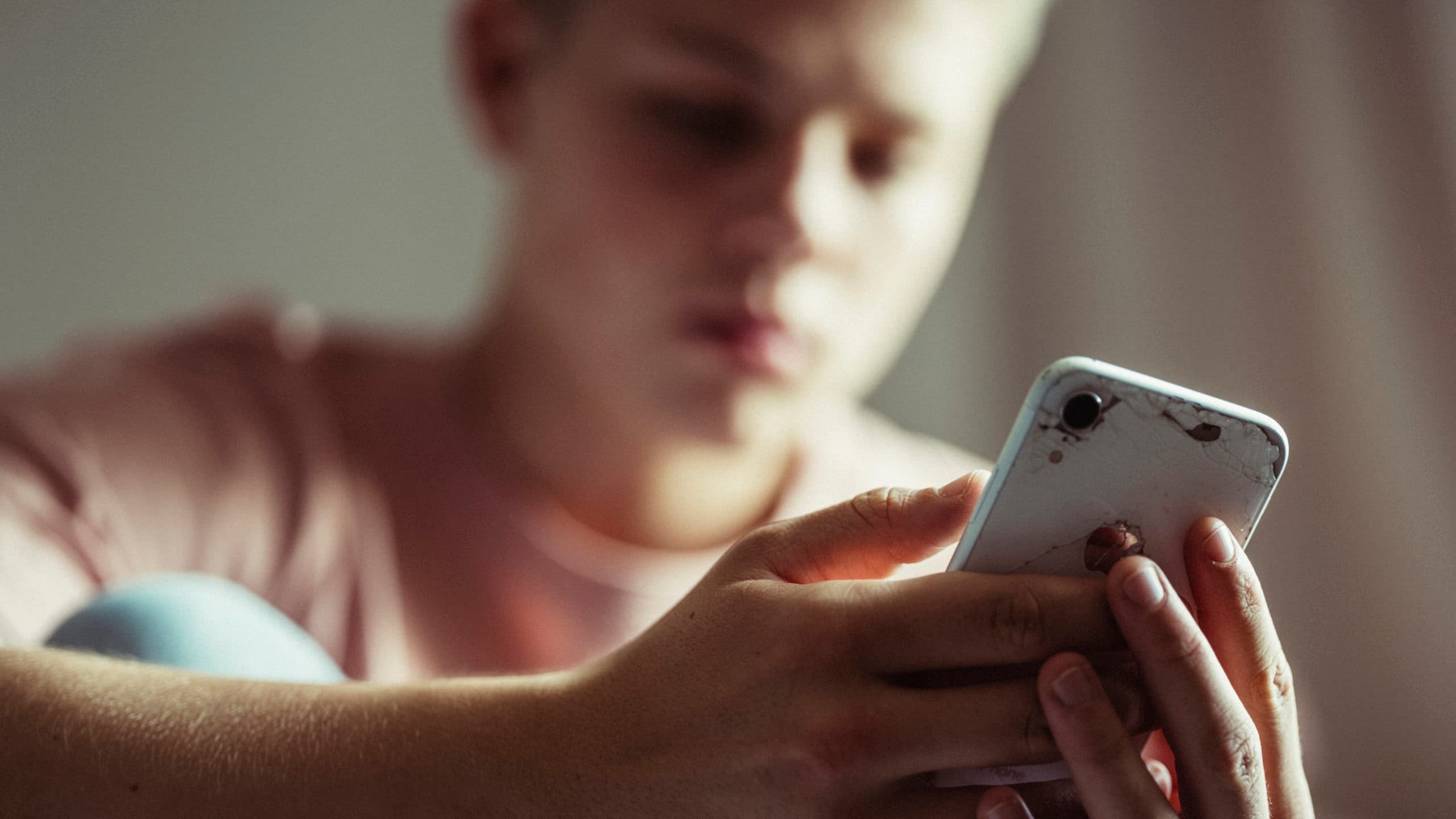 En ung person i rosa skjorte holder og ser på en hvit smarttelefon med sprukket bakside, og ser fokusert ut mens den bruker enheten. Bakgrunnen er svakt uskarp.