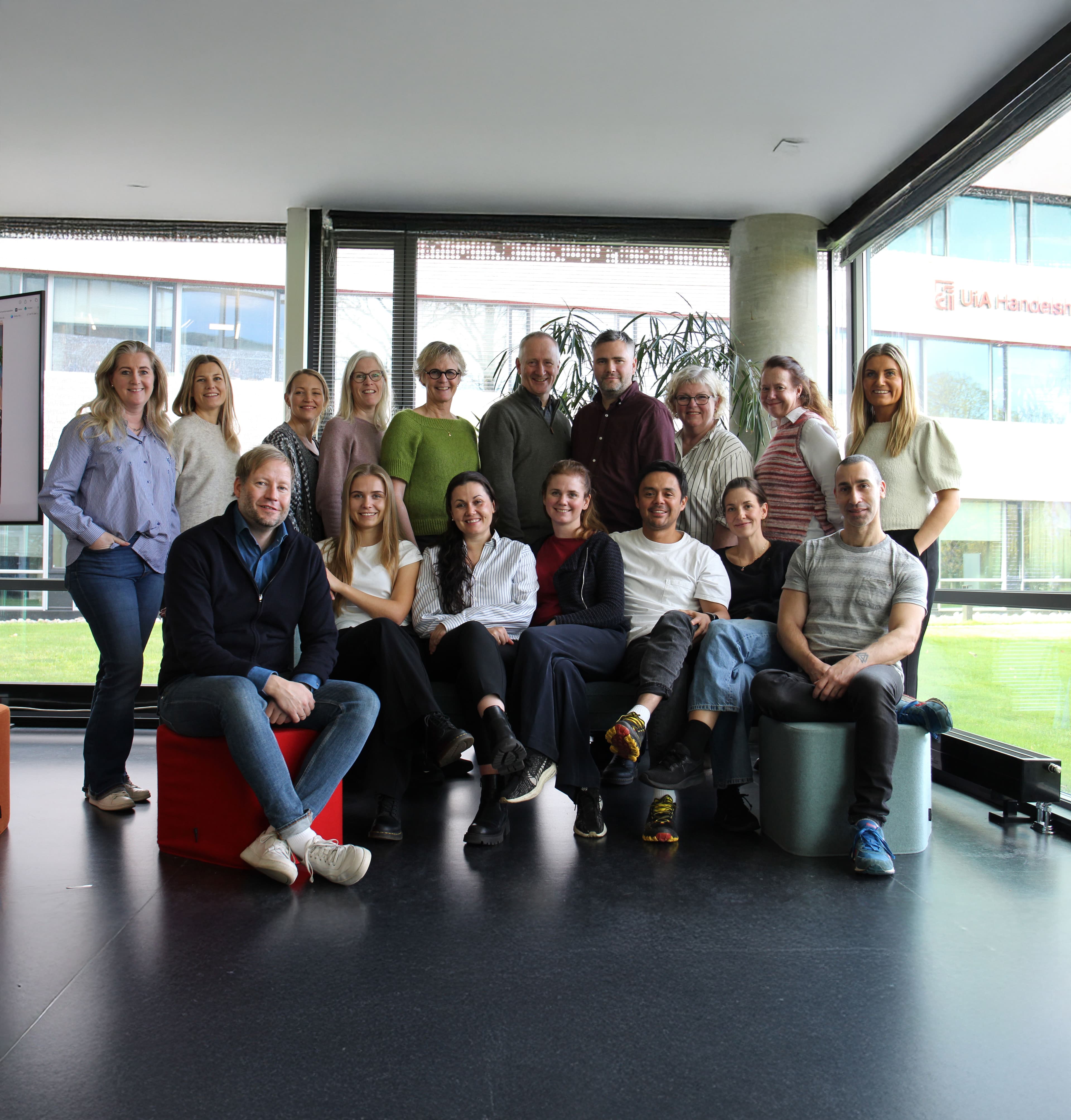 En gruppe på 17 personer, en blanding av menn og kvinner, sitter og står sammen og smiler til et gruppebilde innendørs foran store vinduer med en bygning og grøntområder synlig utenfor.